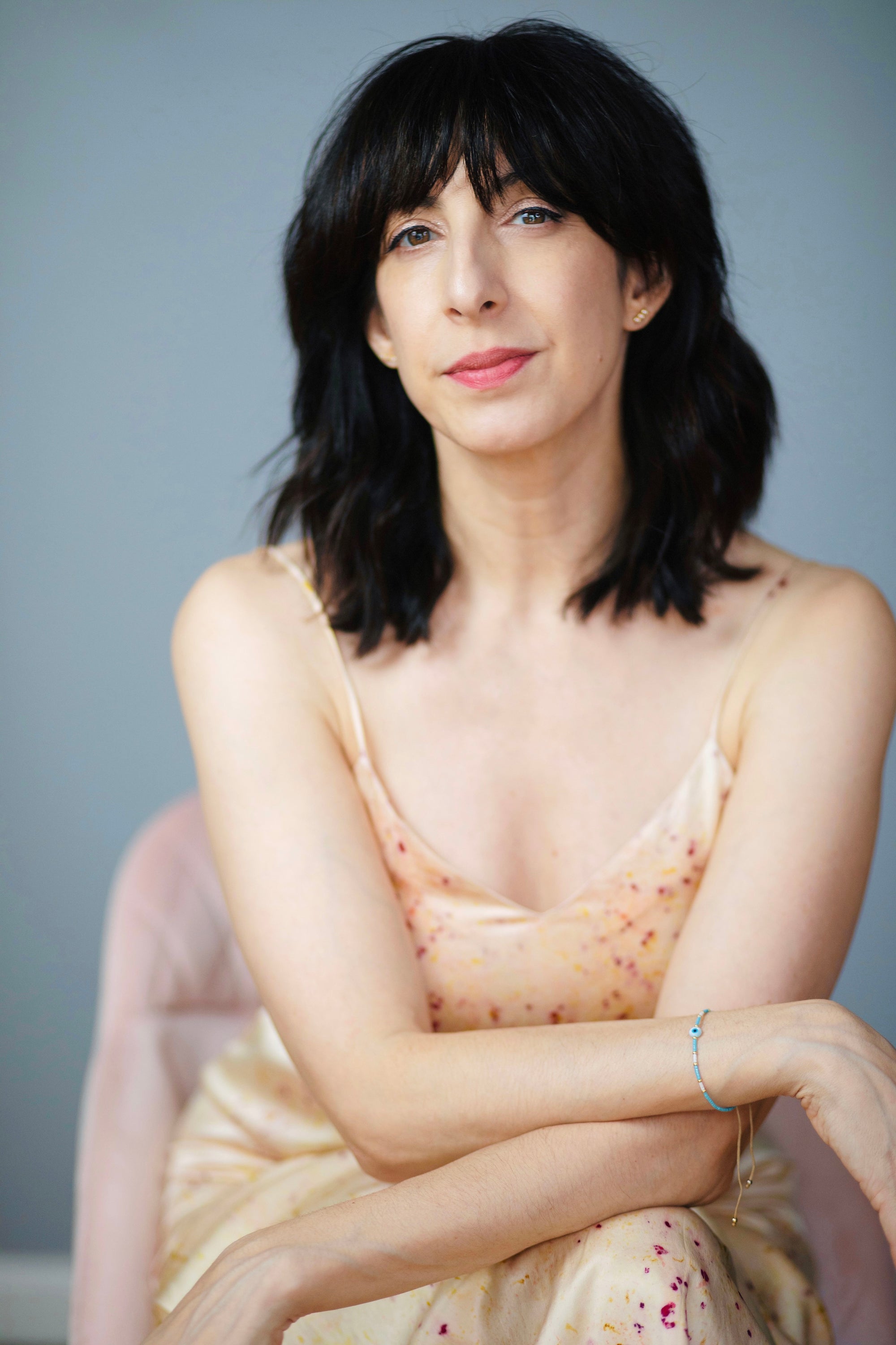 A woman with short dark hair sits against a soft gray background, wearing a pale peach satin camisole with small scattered floral details.